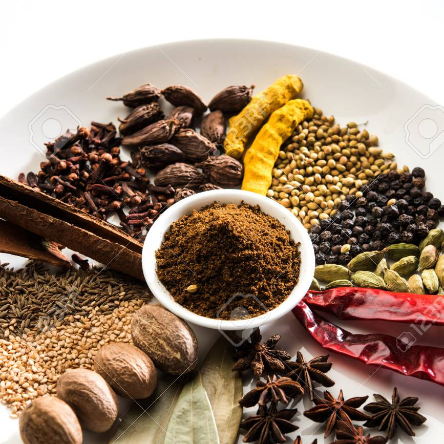 A variety of whole spices and seasonings including cinnamon sticks, cloves, cardamom, cumin, coriander, and whole peppercorns arranged in a bowl on a white plate with a small pile of brown garam masala powder in the center.